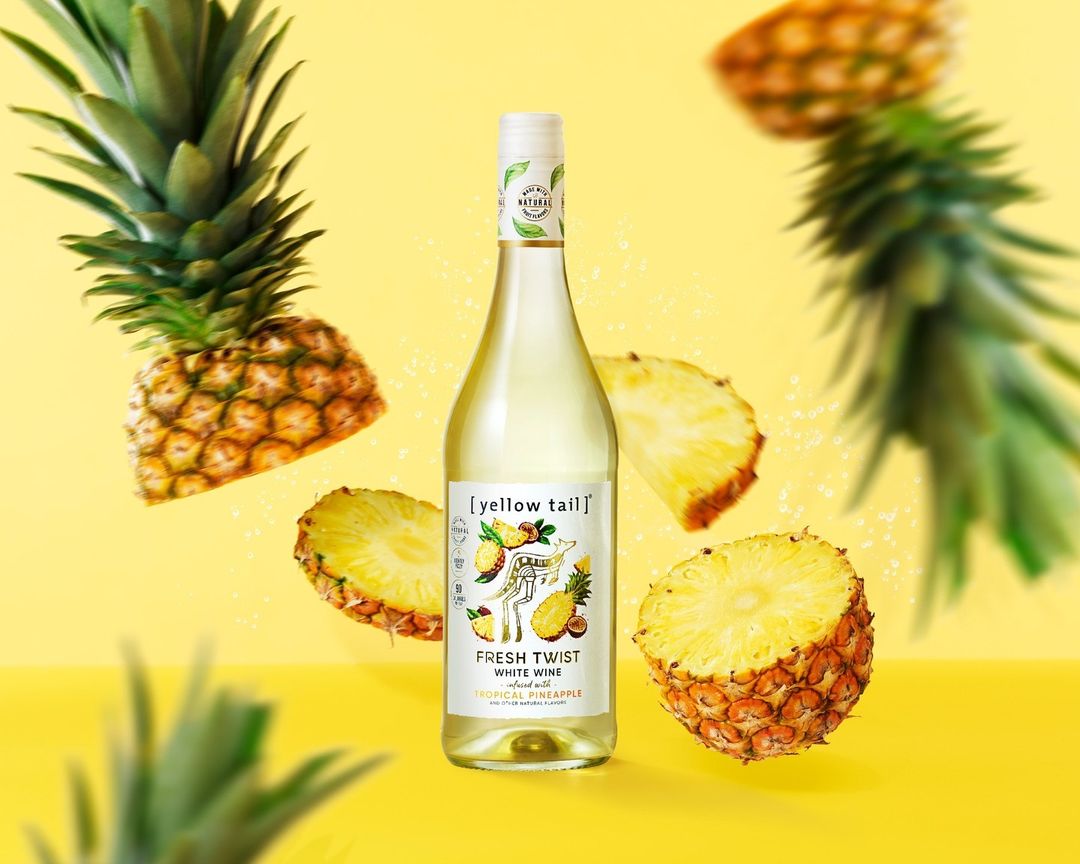 Bottle of Yellow Tail Fresh Twist White Wine with pineapple slice and whole pineapple against a yellow background in a studio shot.