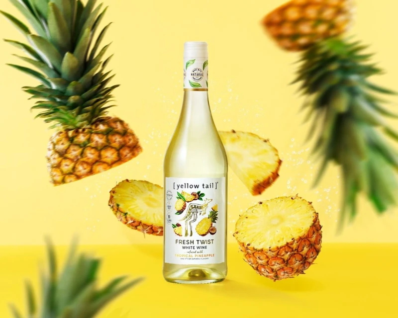 Bottle of Yellow Tail Fresh Twist White Wine with pineapple slice and whole pineapple against a yellow background in a studio shot.