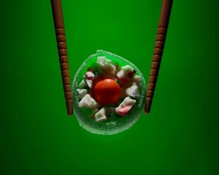 Chopsticks holding a round green candy sushi piece with a central red candy ball against a vibrant green background.
