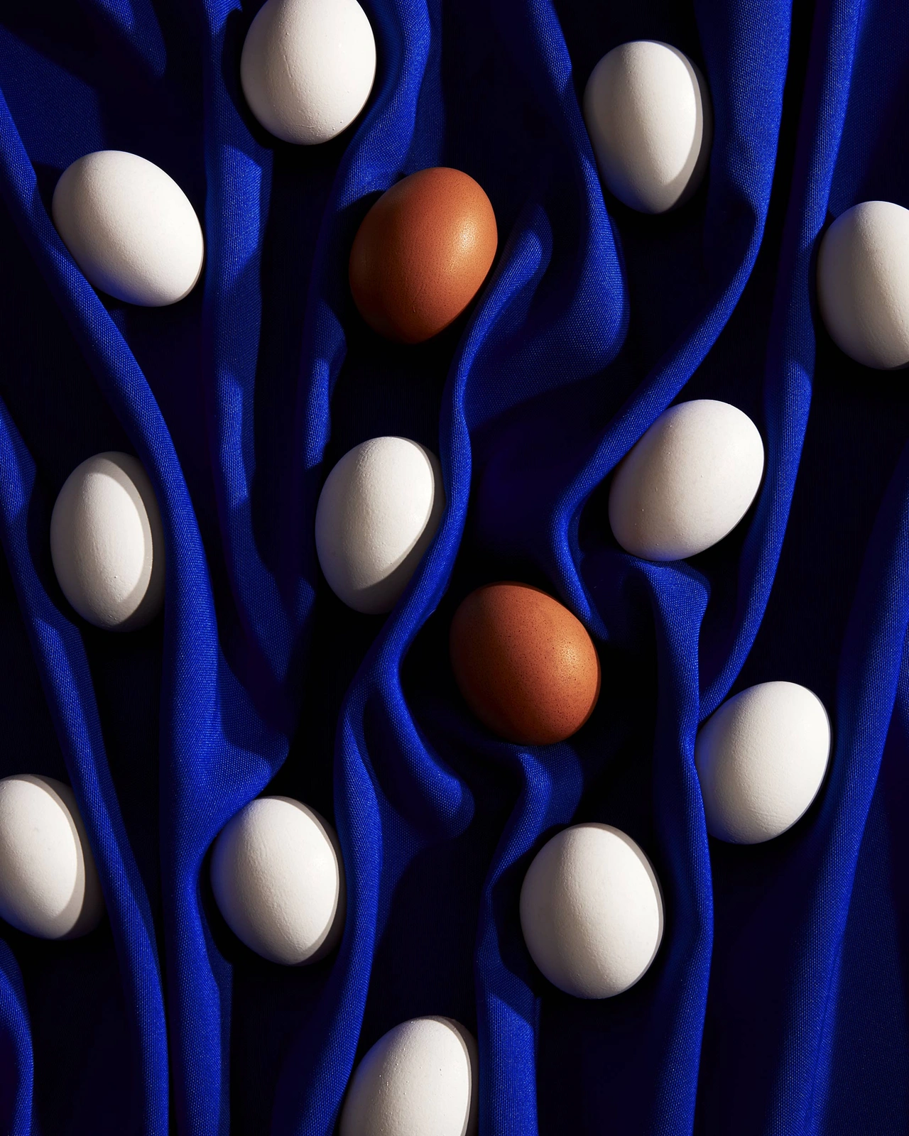 Overhead still life of multiple white eggs and two brown eggs resting on deep blue pleated fabric