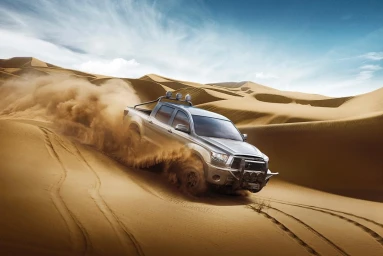 Silver pickup truck driving fast through sand dunes kicking up a thick cloud of dust under a bright blue sky.