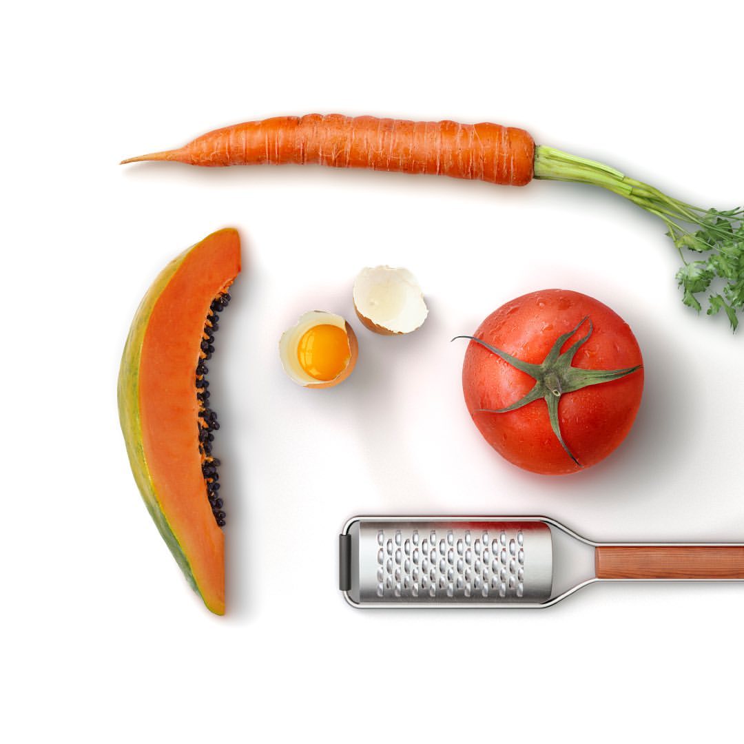 Overhead shot of a sliced papaya, carrot, tomato, raw egg, eggshell and grater on a bright white surface, showcasing fresh food items.
