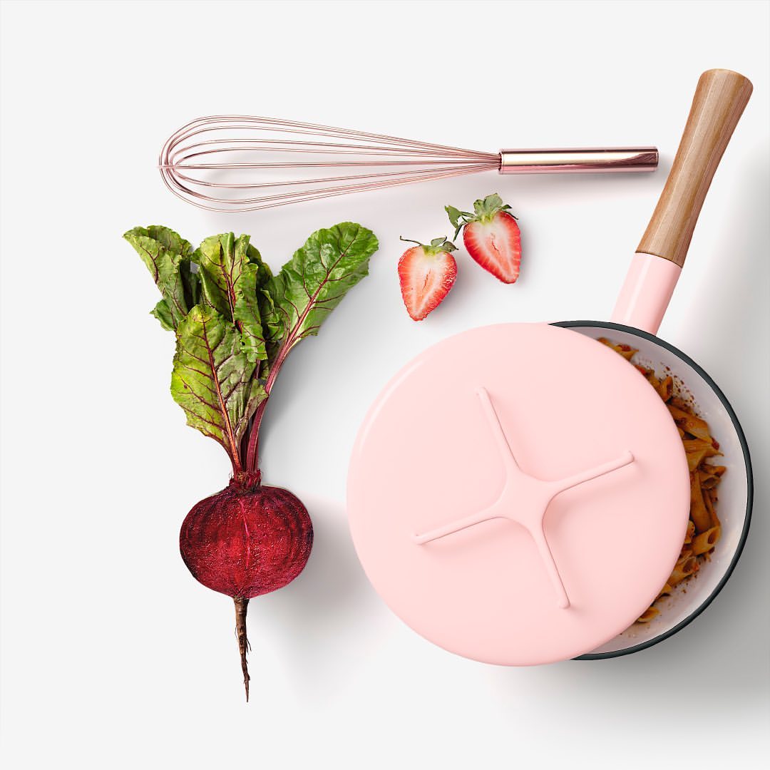 Overhead studio shot: a beetroot, two strawberry halves, a pink-lidded pot filled with pasta, and a whisk arranged on a white surface.