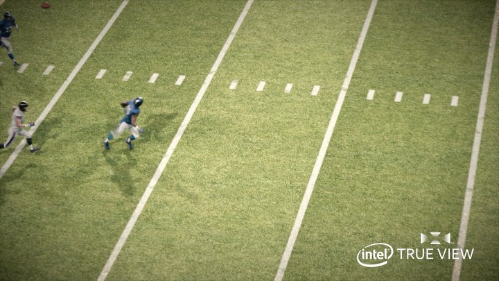 Football players in action on a green field during a game, with white lines marking the field, captured from an overhead perspective.