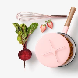 Flat lay of pink saucepan filled with pasta, whole beetroot with greens, two strawberries and a whisk on white