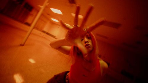 Woman in a red top reaches toward the camera, hand outstretched. Interior setting with fluorescent lighting. Predominantly red color grade.