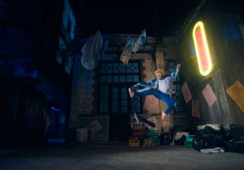 Man in mid-air wearing a blue tracksuit in an alleyway with colorful containers and trash bags and a large rectangular neon sign.