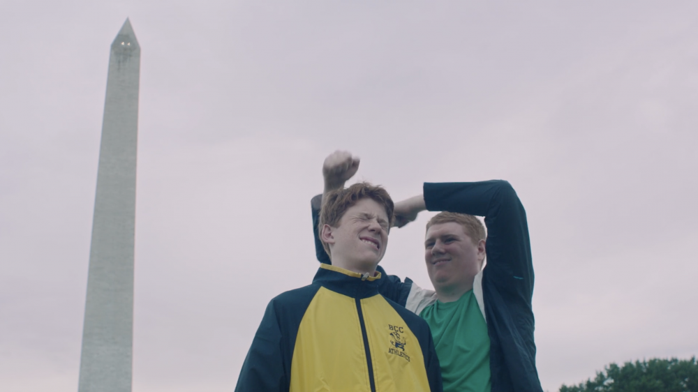 Two young men pose outdoors wearing athletic jackets with the Washington Monument in the background under an overcast sky.