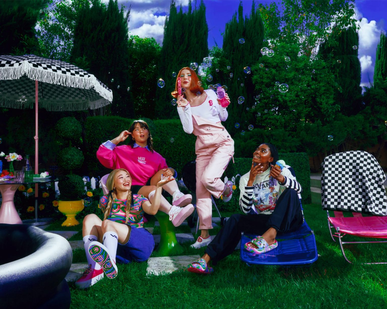 Four young women pose in a garden with bubbles, wearing colorful casual clothes and shoes; green grass, trees, and blue sky background.