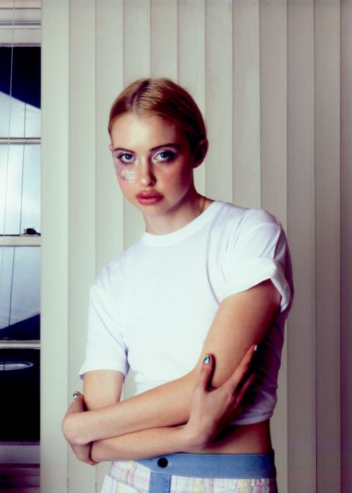 Portrait of a young adult female with blond hair and glitter makeup posing indoors against a neutral vertical-blind background.