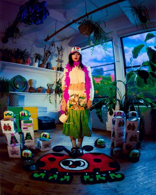 A woman in a creative outfit stands indoors on a ladybug rug surrounded by colorful objects and plants, bathed in diffused light.