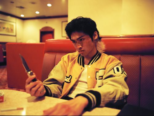 An Asian male sits at a table with a knife and cigarettes in an indoor setting, wearing a yellow jacket with a serious expression.