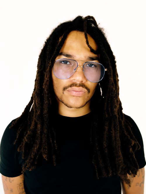 Portrait of an adult male with dreadlocks and glasses, wearing a black t-shirt, against a plain white background in a studio setting.