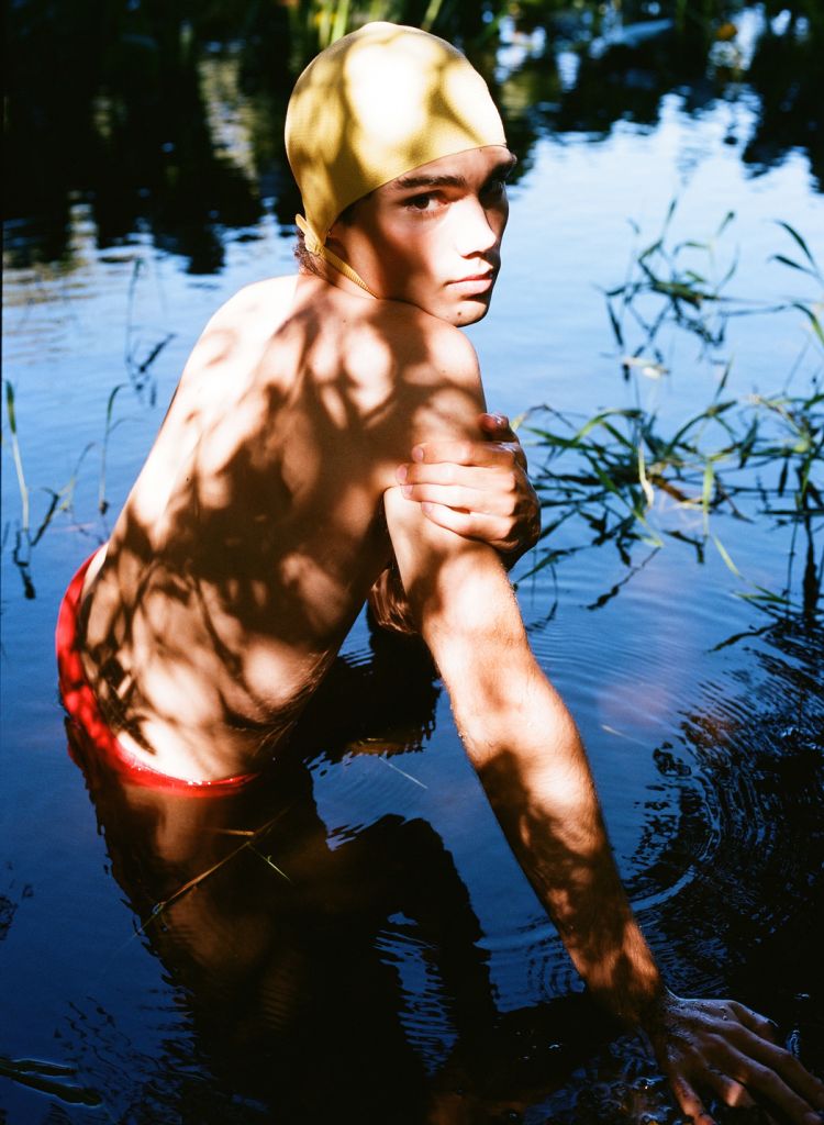 Young shirtless adult male with a yellow swim cap standing in shallow blue water surrounded by tall green grasses and dappled sunlight.