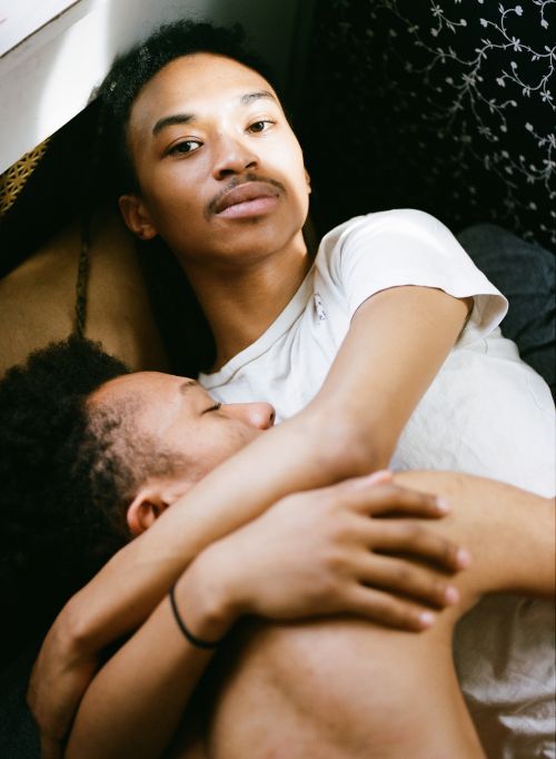 Intimate portrait of two young adult Black men embracing indoors, one shirtless and one in a white t-shirt, set against a patterned curtain.