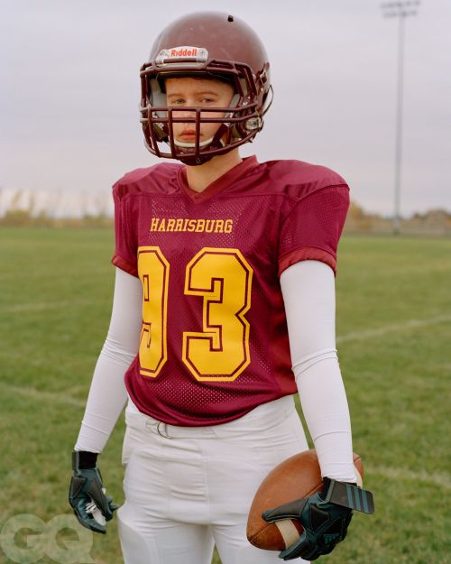 Teenager in football uniform and helmet stands on a field in a medium shot, holding a football on a cloudy day, edited in muted tones.
