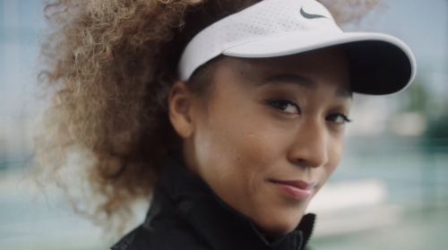 Close-up of Naomi Osaka in a black jacket and white visor, looking at the camera with a slight smile, tennis court in the background.