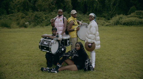 Five diverse individuals outdoors with musical instruments. Three people stand holding snare drums, and two sit in the grassy field.