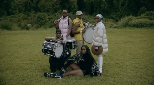Five diverse individuals outdoors with musical instruments. Three people stand holding snare drums, and two sit in the grassy field.