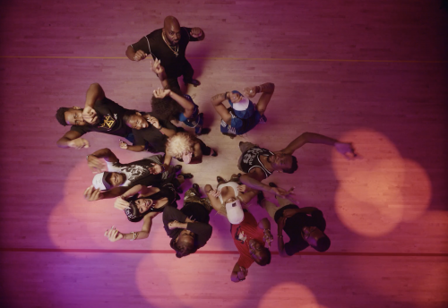 Overhead shot of a group of ten people, including one adult male, posing and dancing in a circle on a hardwood floor with pink lighting.