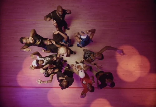 Overhead shot of a group of ten people, including one adult male, posing and dancing in a circle on a hardwood floor with pink lighting.