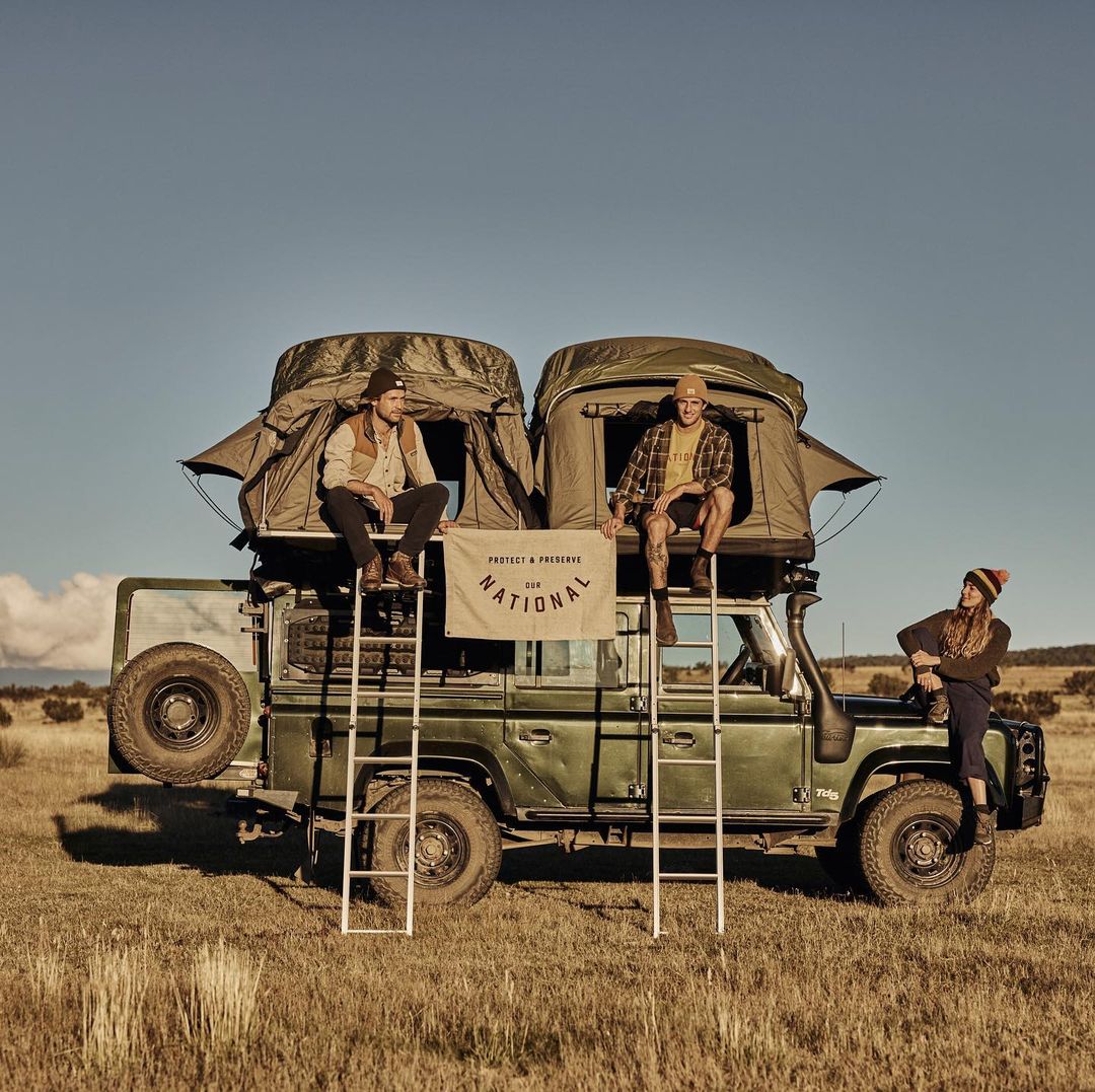 Three people on a green SUV with rooftop tents in a grassy field under a blue sky, enjoying an outdoor adventure.