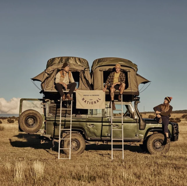 Three people on a green SUV with rooftop tents in a grassy field under a blue sky, enjoying an outdoor adventure.
