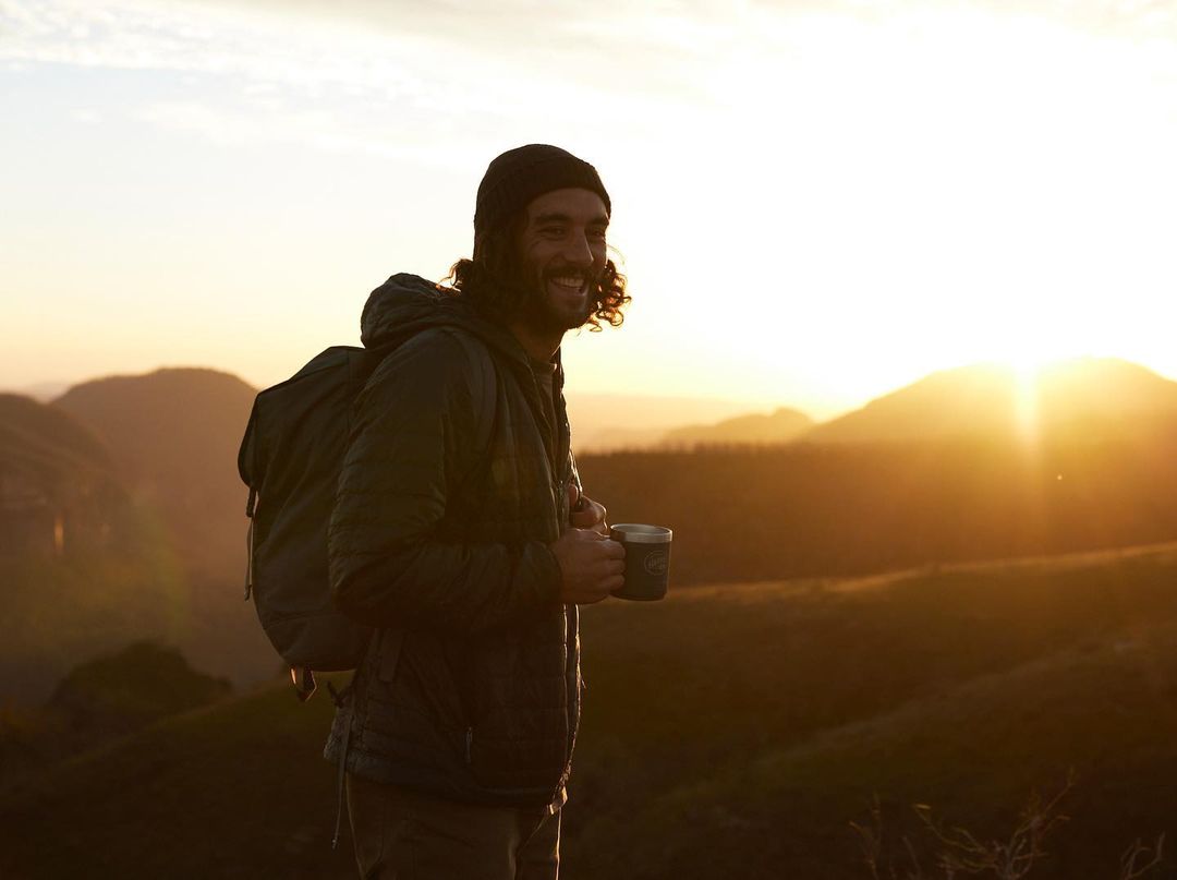 Smiling man with a backpack enjoys the sunrise over rolling hills, holding a mug, wearing a beanie and jacket in a scenic landscape.