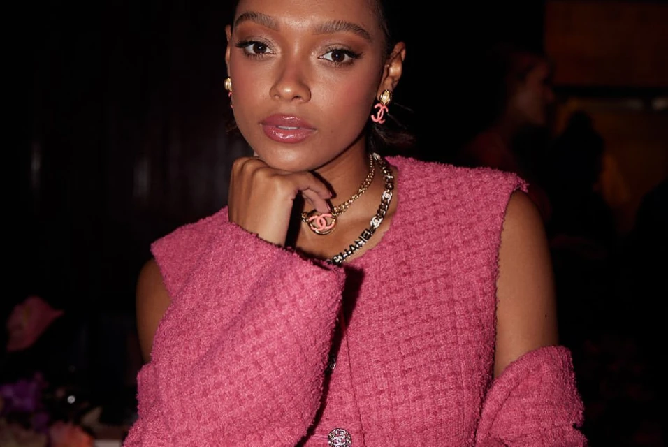 Close-up of a woman wearing a pink tweed vest, gold necklaces, and pink earrings with a hand near her face in a dark indoor setting.