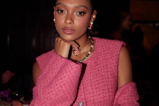 Close-up of a woman wearing a pink tweed vest, gold necklaces, and pink earrings with a hand near her face in a dark indoor setting.