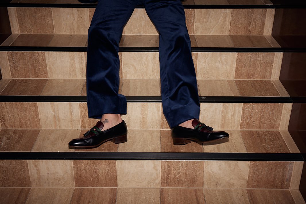 Person in navy velvet pants and black leather tassel loafers standing on a beige and brown tiled staircase with black trim.