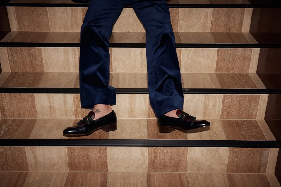 Person in navy velvet pants and black leather tassel loafers standing on a beige and brown tiled staircase with black trim.