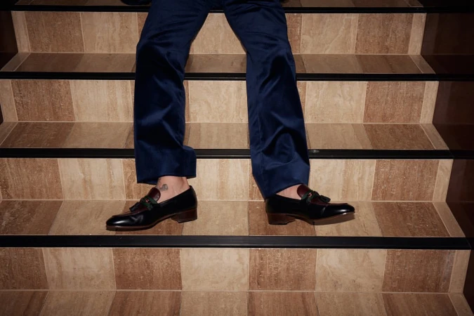Person in navy velvet pants and black leather tassel loafers standing on a beige and brown tiled staircase with black trim.