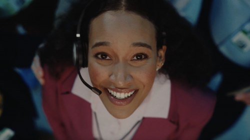 Upward angle of a smiling Black woman wearing a headset and pink blazer, working in a busy office setting with other employees.