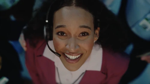 Upward angle of a smiling Black woman wearing a headset and pink blazer, working in a busy office setting with other employees.