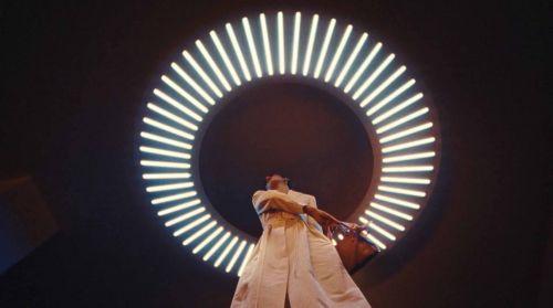 Adult female dancer in a white robe poses underneath a ring-shaped neon light in a dark photo studio with diffused lighting.