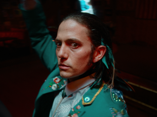 Close-up of a man in a green embroidered jacket with feather hair accessories looking directly at the camera in a dark, indoor setting.