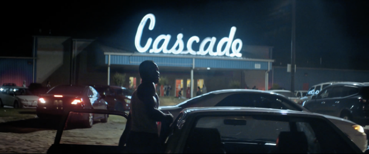 Night scene showing an athletic African American man walking toward a car, with a building lit up with a neon sign behind him.