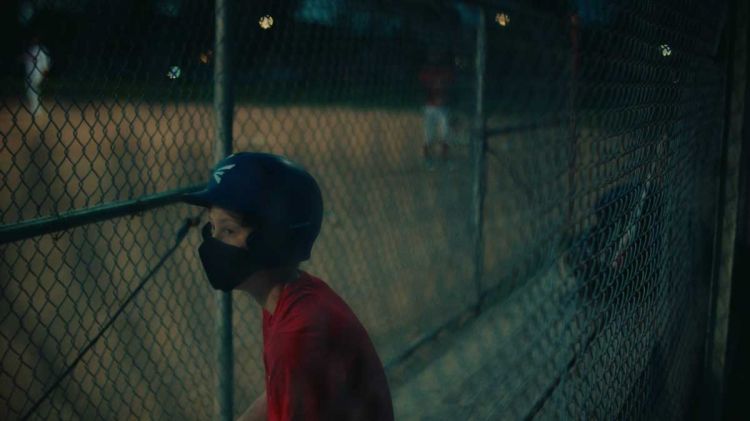 Young baseball player in helmet and face mask stands at a chain-link fence at night, illuminated by dim stadium lighting.