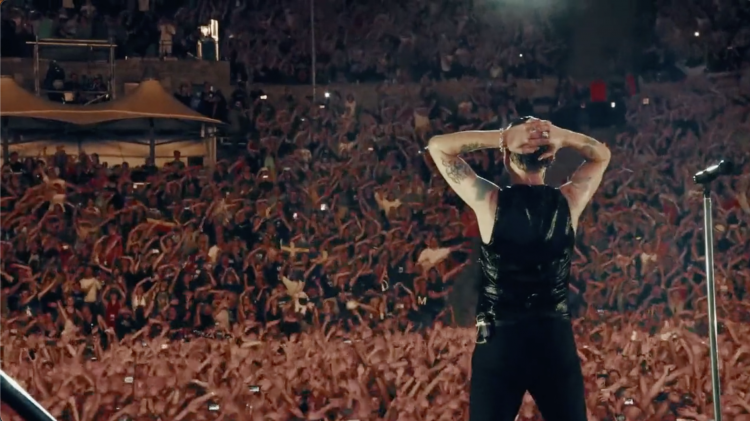 Singer performs on stage with microphone in front of large crowd at concert venue; hands behind head, tattoos visible, dressed in black.