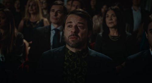 Audience of adults watches an event, featuring an adult male in a patterned shirt and blazer, with surrounding people in formal wear.