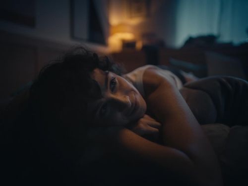 Close-up of an adult woman lying in bed, smiling softly in low light, with dark hair and a light-colored top. Home interior visible.