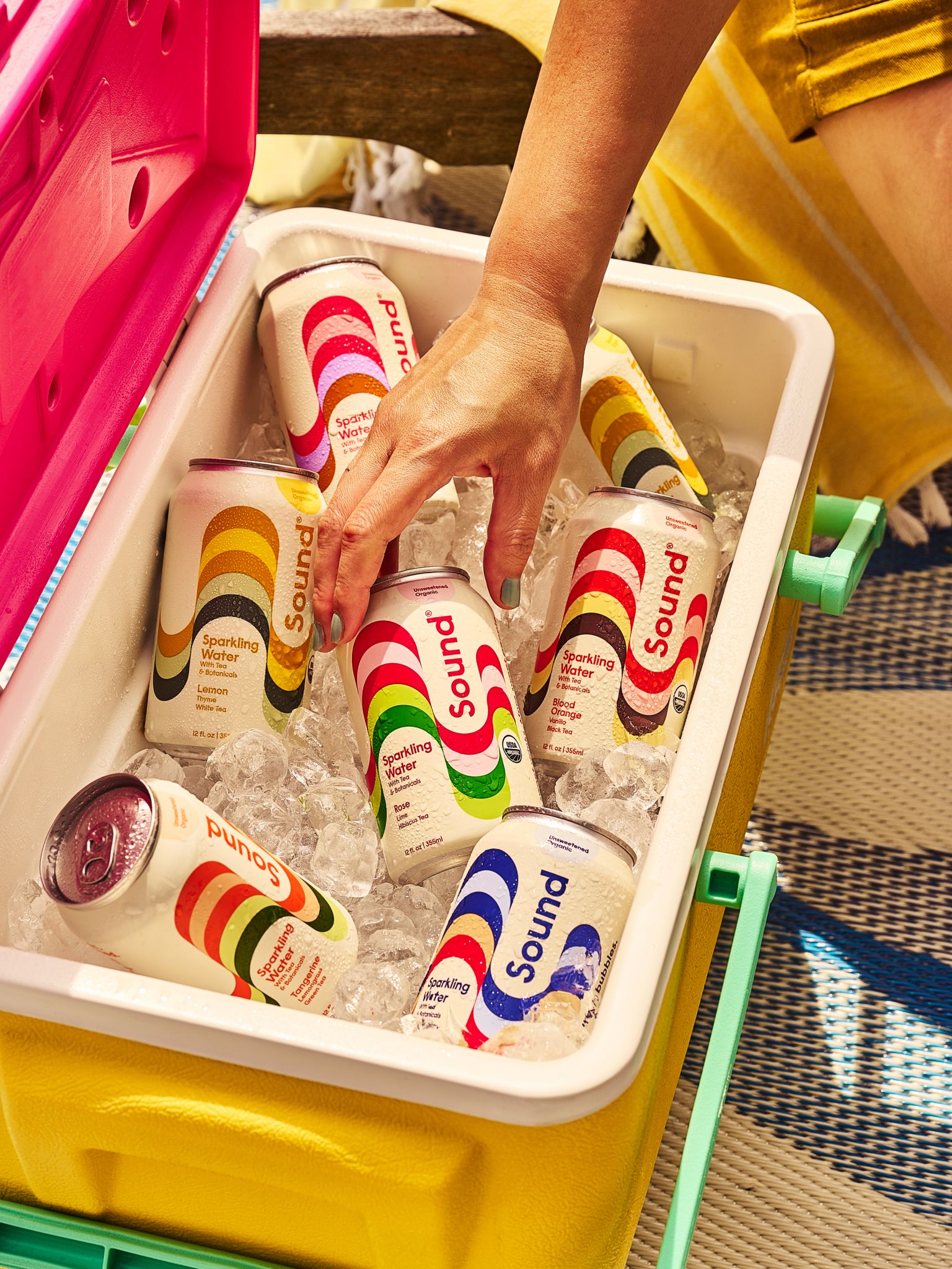 Cooler filled with Sound sparkling water cans in various flavors, hand reaching in, inviting and refreshing, summertime picnic vibe.