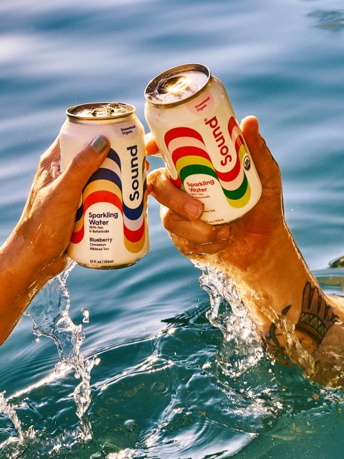 Two cans of Sound Sparkling Water are being held above the water, splashing with some of the water visible in the background.