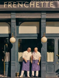 Two male chefs in white shirts and blue aprons standing outside the Frenchette restaurant entrance photographed by Louise Palmberg