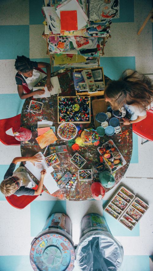 Overhead shot of three children at a round table in an art studio, drawing and using art supplies; the floor is tile.