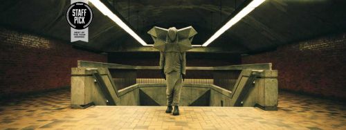 Person in a futuristic, geometric-shaped costume walks away from the camera towards a subway entrance with artificial lighting.