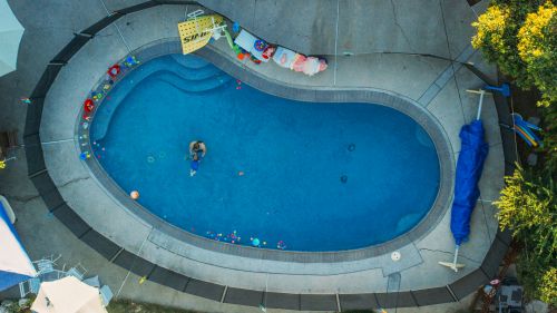 Aerial view of a person in a pool surrounded by toys, with trees and patio visible. The pool has bright blue water.