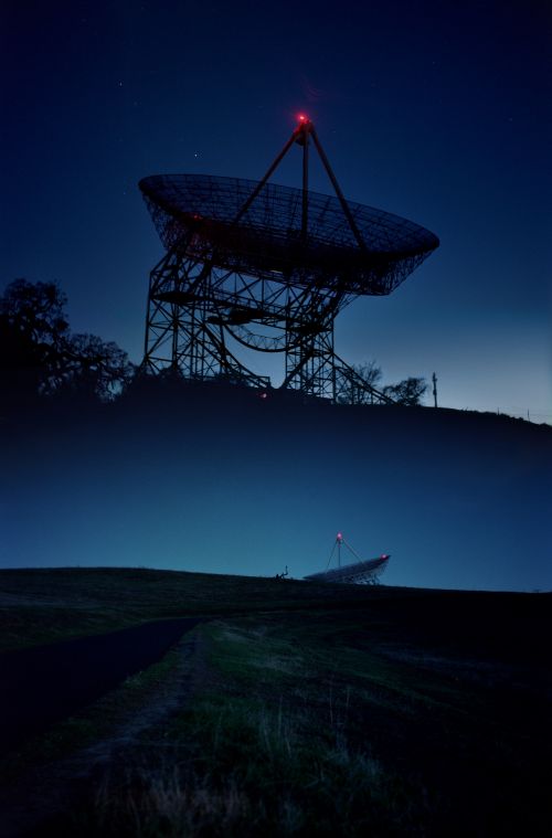 Dramatic low-angle shot of two large radio telescopes against a dark blue sky, showcasing their intricate structures on grassy hills.