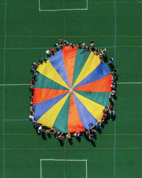 Aerial view of a large group of people holding a colorful parachute-like canopy on a green surface with white lines visible.